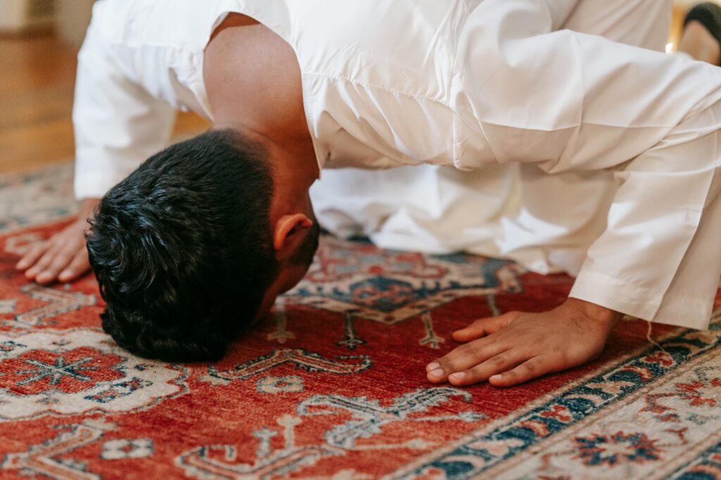 A man in traditional attire kneels in prayer on an elaborate rug at home, embodying cultural and religious devotion.